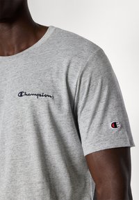 Grå bomulds t-shirt med korte ærmer, der har en navy broderet "Champion" logo på venstre bryst og et lille farveaccent logo på ærmet.