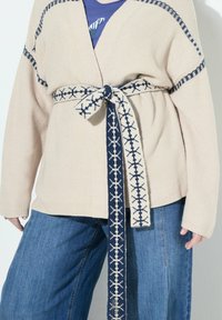 Beige texturerad cardigan med långa ärmar, kompletterad med ett mönstrat marinblått bälte med vita geometriska mönster. Bärs över blå vida jeans.