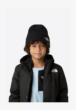 Čierna rebrovaná pletená čiapka s logom, ktorú nosí dieťa. Dieťa má na sebe čiernu bundu s farebnými blokmi v svetlomodrej a čiernej farbe na hornej časti.