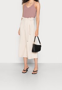 Haut rose pâle sans manches, culottes crème taille haute avec ceinture nouée, et sac à main noir. Des sandales noires à brides complètent la tenue.
