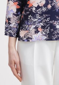 Top navale a fantasia floreale con maniche a tre quarti, caratterizzato da intricati fiori nei toni del rosa, viola e bianco, abbinato a pantaloni bianchi.