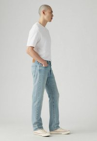 Helle blaue Jeans mit geradem Schnitt, kombiniert mit einem weißen T-Shirt mit kurzen Ärmeln und beigen Sneakers. Mit einem braunen Ledertag.