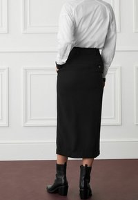 Schwarzer Midirock mit schmaler Taille, Rückentaschendetail und subtiler Textur, kombiniert mit schwarzen, hochhackigen Stiefeletten und einem weißen Hemd.