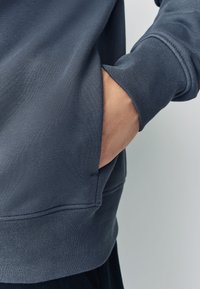 Grijze crewneck-trui met geribde manchetten, zak aan de zijkant en een gladde stoftextuur. Mouw reikt net voorbij de pols.