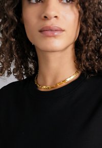 Colier de aur cu zale alungite, așezat pe un hanorac negru cu guler rotund. Părul creț încadrează linia gâtului, subliniind bijuteria.
