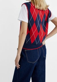 Marineblauwe en rode argyle gebreide mouwloze trui met witte details, gedragen over een wit T-shirt, gecombineerd met donkere denim broek.