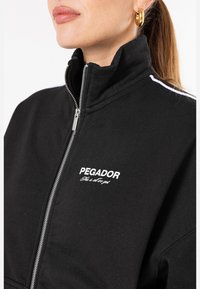 Čierna zateplená bunda na zips z bavlny, s vysokým golierom, bielym logom "PEGADOR" na hrudi a bielymi pruhmi pozdĺž švov na pleciach.
