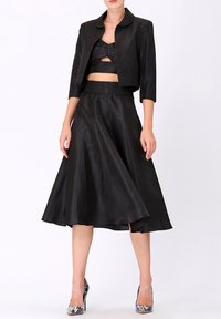 Veste noire en satin coupée avec un col arrondi, associée à une jupe noire évasée assortie. Le mannequin porte des chaussures à talons colorées.