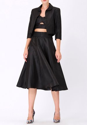 Veste noire en satin coupée avec un col arrondi, associée à une jupe noire évasée assortie. Le mannequin porte des chaussures à talons colorées.