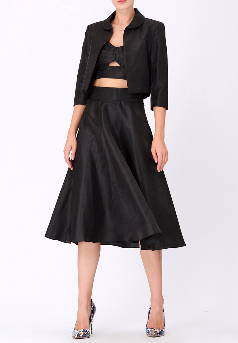 Veste noire en satin coupée avec un col arrondi, associée à une jupe noire évasée assortie. Le mannequin porte des chaussures à talons colorées.