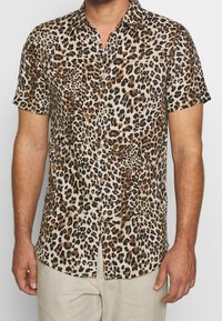 Mand iført en kortærmet skjorte med brun og sort leopardprint, sammen med lyse beige bukser.