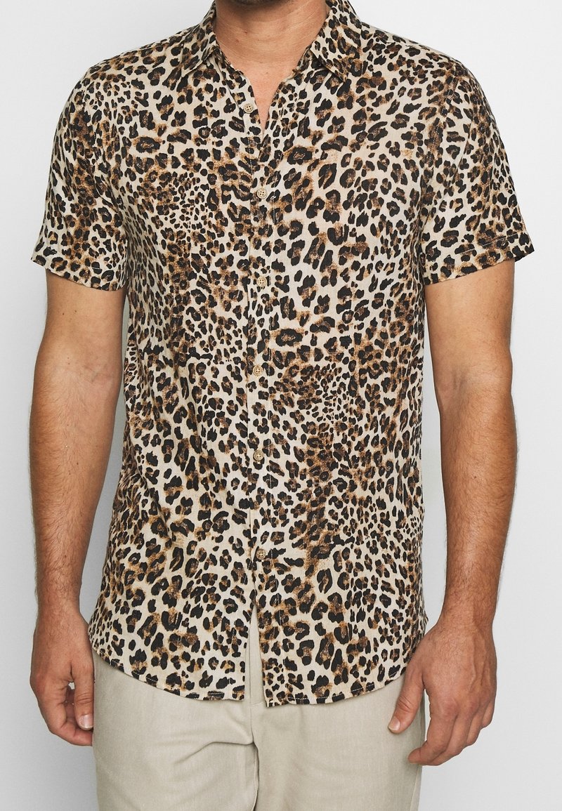 Mand iført en kortærmet skjorte med brun og sort leopardprint, sammen med lyse beige bukser.