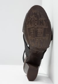 Semelle d'une chaussure à talon noire avec un texte en relief "WE WANT FELMINI" et le numéro 37, montrée sur un fond blanc.