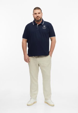 Man with beard wearing navy polo shirt, beige trousers, and white sneakers standing with one hand in pocket on white background.