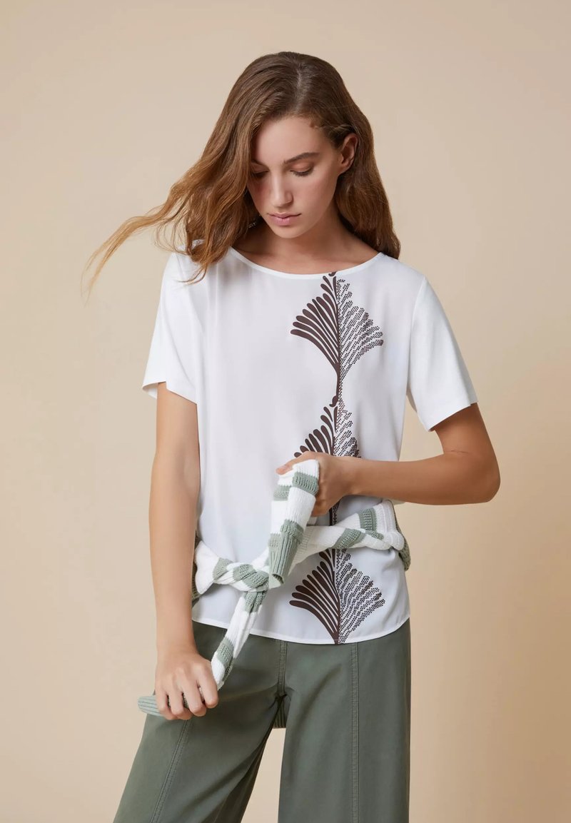 T-shirt bianco a maniche corte con un motivo a foglia marrone scuro davanti, abbinato a pantaloni verde oliva. Un maglione a righe crema e verde legato in vita.