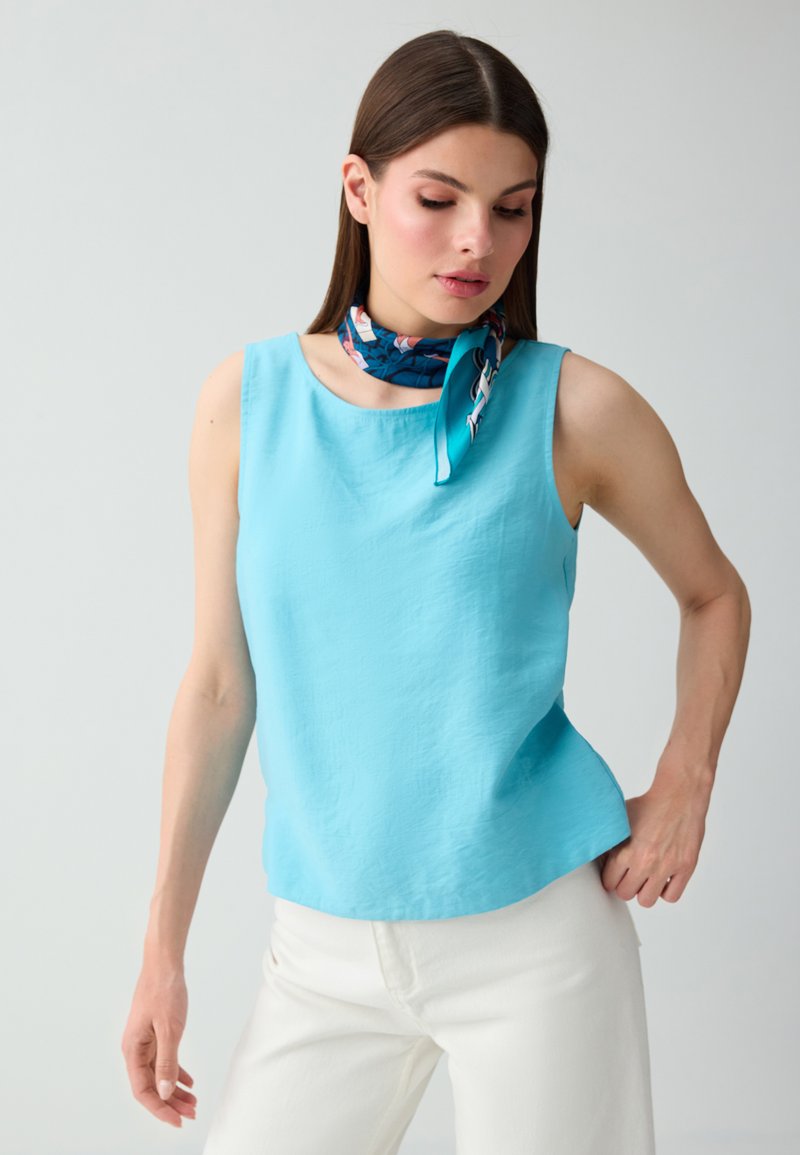 Femme aux cheveux bruns et raides portant un haut sans manches bleu clair, un pantalon blanc et un foulard bleu foncé à motifs autour du cou, regardant vers le bas.