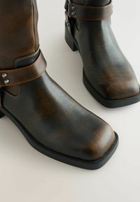 Brązowe skórzane botki za kostkę o teksturowanym wykończeniu, z kwadratowym noskiem i czarną gumową podeszwą. Posiadają regulowany pasek i srebrne ćwieki.