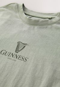 Zelena bombažna majica s posadkim ovratnikom. Na sprednji strani je črn natisnjen logotip harfe in napis "GUINNESS ESTD 1759". Mehka tekstura.