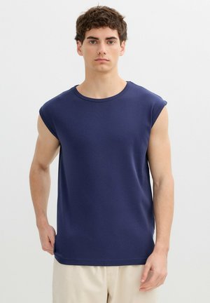 Joven con cabello corto, rizado y castaño, vistiendo una camiseta sin mangas azul marino y pantalones beige, de pie sobre un fondo blanco liso.