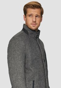 Grijze herringbone jas met een hoge kraag, voorzien van zwarte accenten en een ritszak. Getextureerde stof en een getailleerde pasvorm.