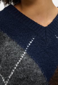 Gros plan d’une personne portant un pull en maille texturée avec des blocs de couleur bleu marine, gris et marron, et des détails de points blancs près de l’encolure.