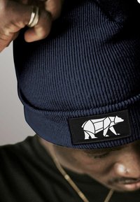 Person wearing a dark blue knit beanie with a geometric white bear patch, adjusting the hat with a hand wearing a silver ring.