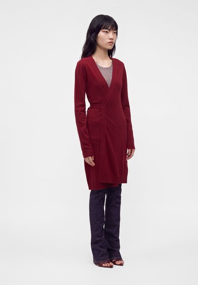 Femme portant un long pull cache-cœur rouge sur un haut beige, un pantalon évasé foncé et des talons ouverts, debout devant un fond uni.