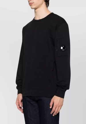 Giovane uomo che indossa una felpa nera a maniche lunghe con una tasca con cerniera e una toppa circolare sulla parte superiore della manica sinistra, abbinata a jeans scuri.