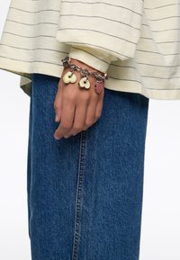 Pulsera de cadena de plata con tres charms en forma de corazón de colores: verde, crema y rojo, llevada en una mano con una manga de rayas amarillas.