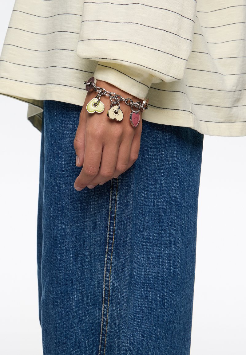 Pulsera de cadena de plata con tres charms en forma de corazón de colores: verde, crema y rojo, llevada en una mano con una manga de rayas amarillas.