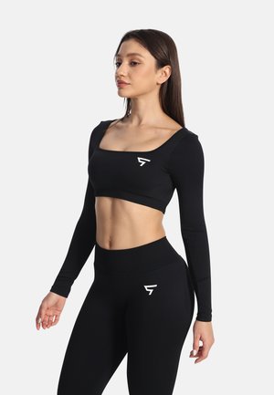 Kobieta w czarnej, długorękawowej bluzce typu crop top oraz legginsach z wysokim stanem z białymi logo, stojąca z rozluźnionymi ramionami na jednolitym tle.