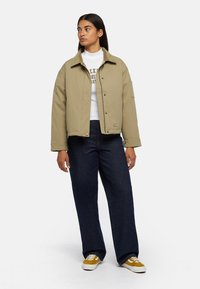 Giacca khaki chiaro con colletto morbido, bottoni a scatto e logo a patch, abbinata a jeans a gamba larga blu scuro e sneakers gialle.