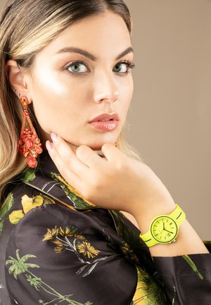 Reloj de silicona amarillo brillante con esfera redonda en la muñeca, combinado con grandes pendientes florales naranjas. El fondo es de un beige suave y la camisa tiene un estampado floral.