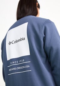 Modrá mikina s velkým bílým čtvercem na zadní straně se znakem "Columbia" a textem: "SINCE 1938 PORTLAND, OREGON, USA."