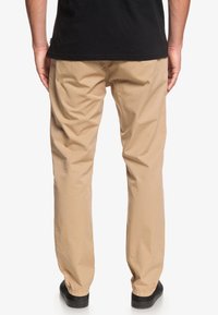 Ljusbeige chinos med lös passform, med en slät textur och elastiska muddar. Bärs med en svart skjorta, med minimala detaljer synliga.