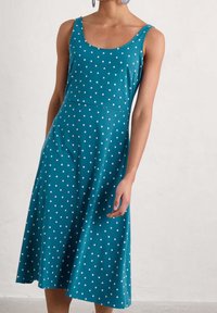 Tealfärgad ärmlös klänning med vita prickar, figursydd överdel, utsvungen kjol och mjuk textur. Har en rundad halsringning och sidofickor.