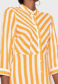 Camisa a rayas en colores naranja y blanco, con botones en el frente, collar y un bolsillo en el pecho con un patrón de rayas diagonales. Tejido suave, de mangas largas.