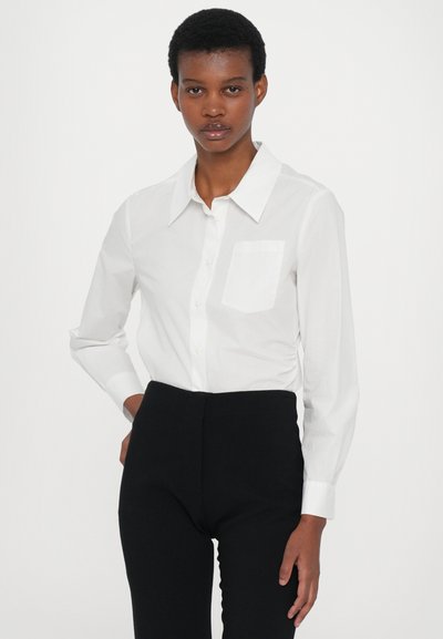 Witte button-up shirt van gladde stof, met een puntige kraag, lange mouwen en een enkele borstzak. Gecombineerd met strakke zwarte broek.
