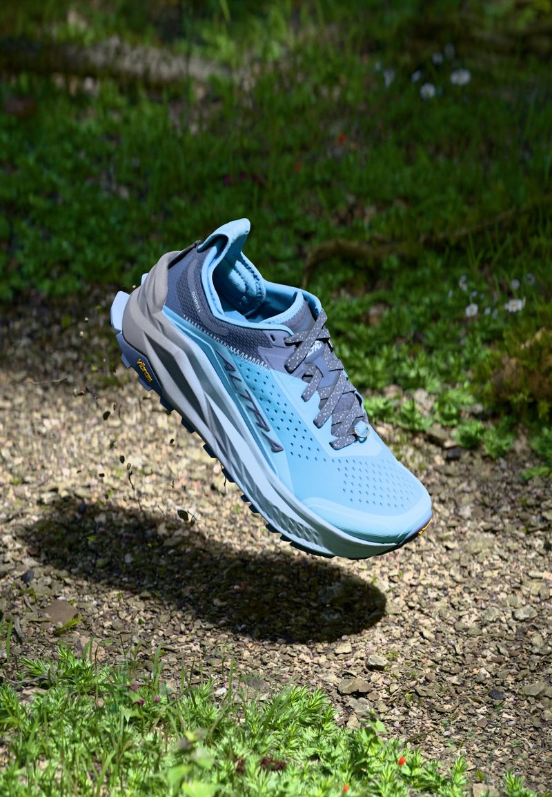 Baskets de trail de couleur bleu clair avec des accents gris, dessus en mesh respirant, semelle texturée et système de laçage proéminent, flottant au-dessus du sol.