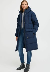 Navy blue puffer coat with a hood and front zip, featuring two side pockets and one chest pocket, worn with fitted jeans and ankle boots.