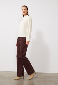 Haut à manches longues crémeux avec col montant, associé à un pantalon en velours côtelé bordeaux à jambes larges et des escarpins beiges à bout pointu, le tout sur fond blanc.