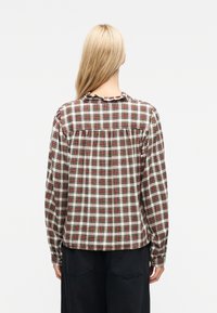 Γυναίκα με μακριά ξανθά μαλλιά, γυρισμένη με την πλάτη, φορώντας μια μακρυμάνικη κόκκινη, λευκή και μαύρη καρό πουκάμισο και σκούρο παντελόνι σε λευκό φόντο.