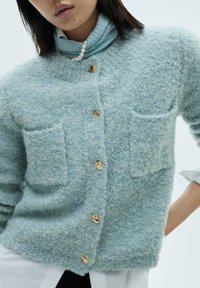 Cardigan azzurro chiaro e morbido con due tasche frontali e bottoni dorati. Indossato sopra un dolcevita azzurro chiaro, presenta una texture delicata.