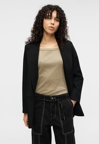 Μαύρο σακάκι με πέτο, πάνω από μπεζ αμάνικο τοπ. Μαύρο παντελόνι με λευκές ραφές και ζώνη στη μέση με κορδόνι. Σγουρά μαλλιά που πλαισιώνουν το πρόσωπο.