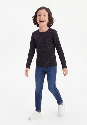 Smilende barn med skulderlangt hår iført sort prikket langærmet skjorte, blå jeans og hvide sko går fremad mod hvid baggrund.