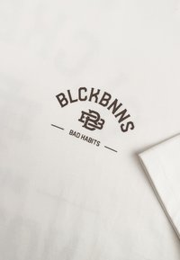 Witte katoen T-shirt met bruine bedrukte tekst: "BLCKBNNS," "PB3," en "BAD HABITS." Soepele textuur en standaard ronde halsontwerp.