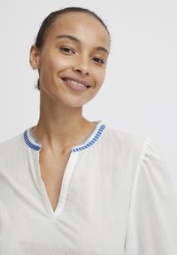 Blouse blanche avec un col brodé bleu, une ouverture à patte et des manches bouffantes. Le tissu semble léger et texturé.