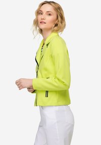 Limoengroene jas met zwarte accenten, slim fit, met ritszakken en een reverskraag. Gedingd over witte broek, wat een soepele textuur laat zien.