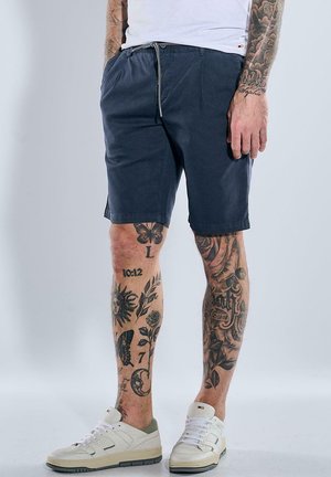 Navy blue cotton shorts with a drawstring waist, side pockets, and a tailored fit, paired with white sneakers and tattooed legs.