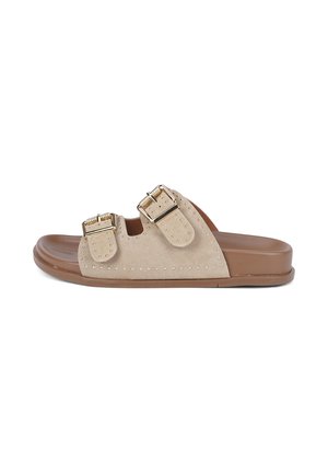 Beige Wildleder-Pantoletten mit zwei verstellbaren goldenen Schnallenriemen und kleinen Metallnieten auf einer braunen flachen Sohle.